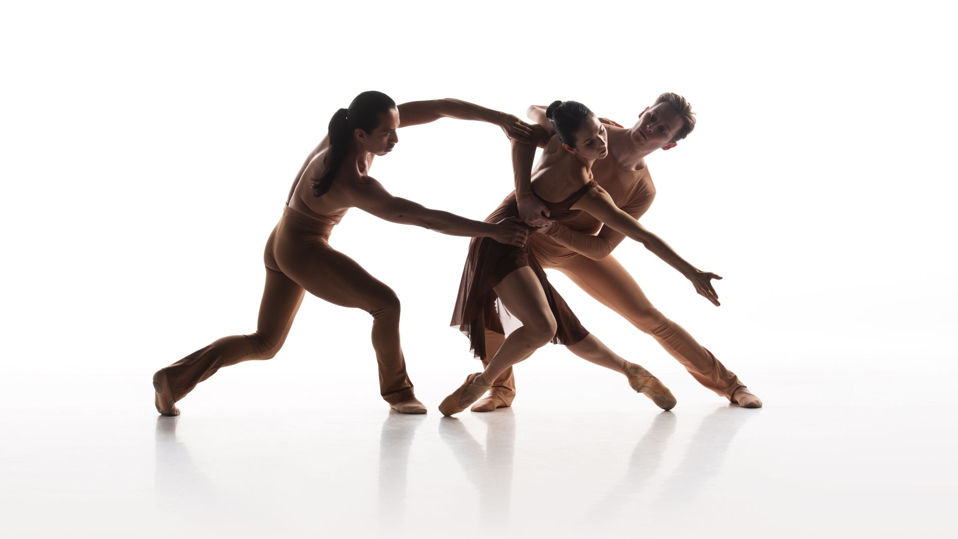 Three dancers in skin-toned outfits. One holding another and one reaching out to the other.