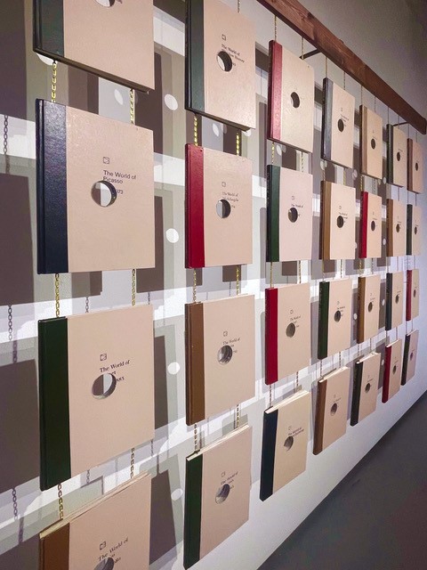 Books hanging from block of wood connected by chains. Each book has a hole missing from the centre.